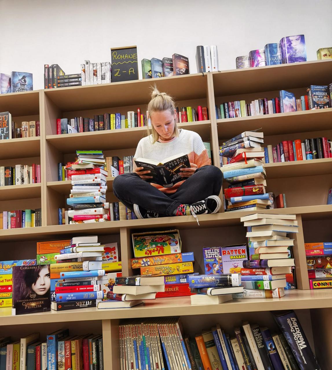Frau sitzt im Schneidersitz zwischen Büchertürmen und Bücherregalen